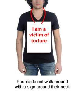 Image of a man in jeans and a black t-shirt, with a big sign around his neck with red writing saying 'I am a victim of torture' 